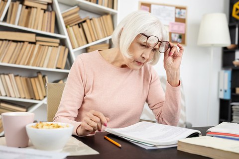 Eine ältere Dame sitzt mit konzentriertem Blick an einem Arbeitstisch und liest in einem Dokument.